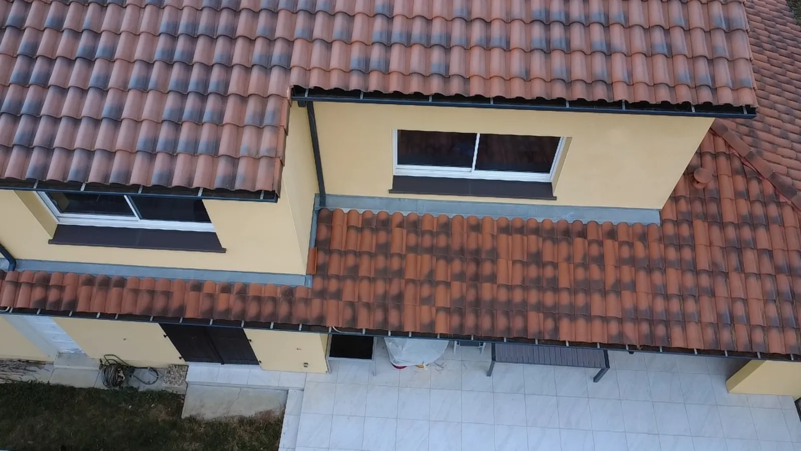 Vue oblique par drone de la jonction entre deux pans de toiture en tuiles terre cuite patinées, avec gouttière et fenêtres de façade