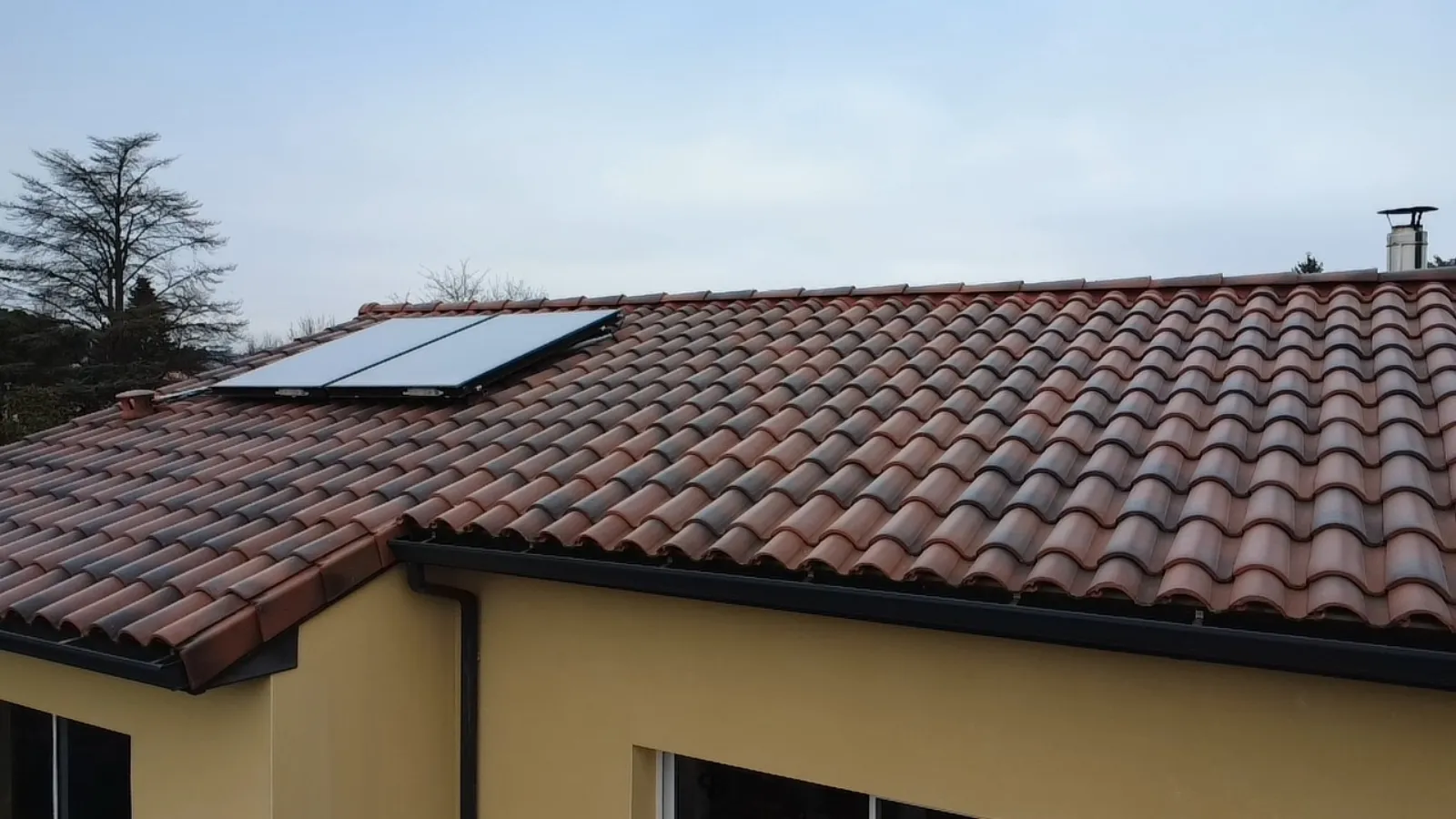 Pan de toiture en tuiles terre cuite avec deux panneaux solaires thermiques et souche de cheminée, façade jaune en contrebas