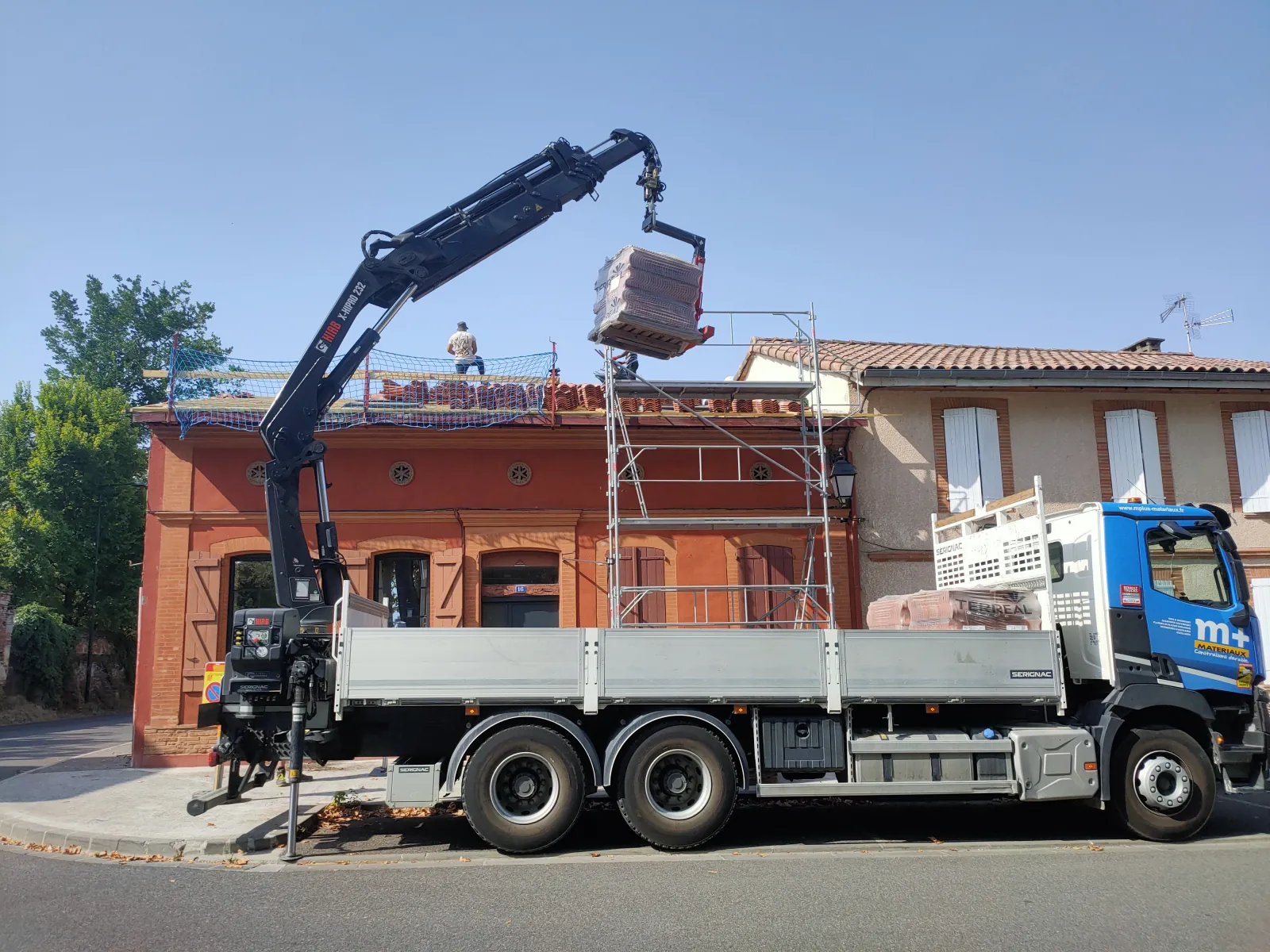 Camion-grue levant une palette de tuiles vers le toit d'une maison ancienne en briques en cours de rénovation de couverture